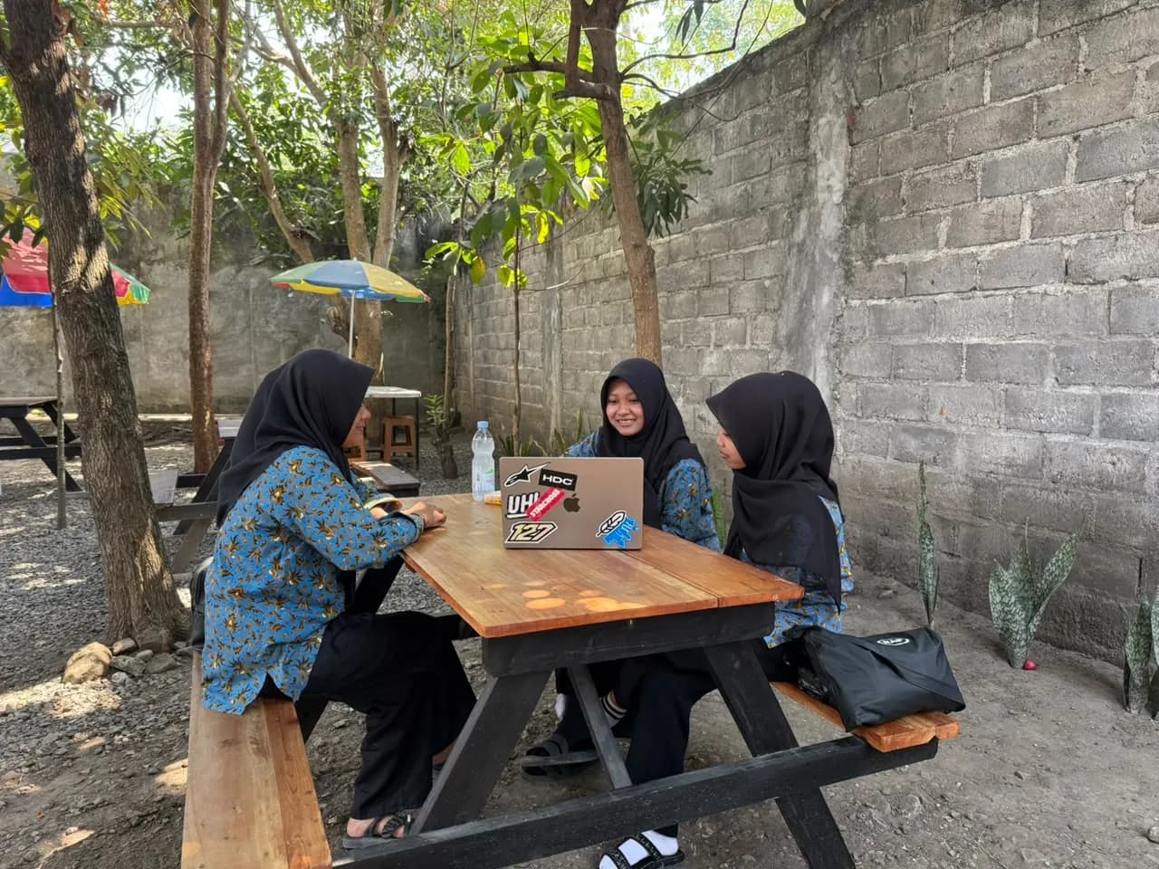 Tiga peserta bekerja dengan laptop di meja kayu area terbuka